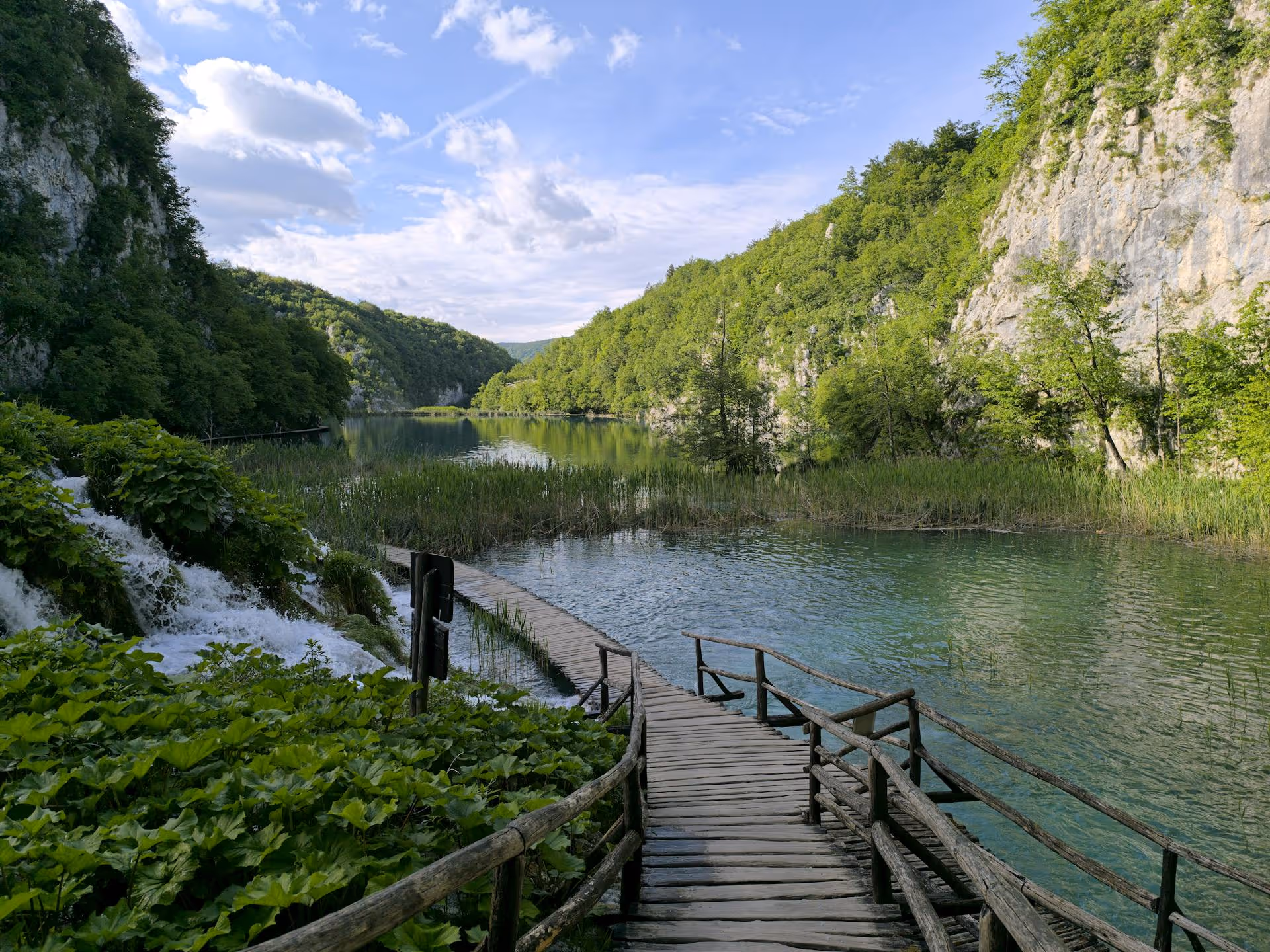 A rustic boardwalk leading across a lake flanked by lush mountains.