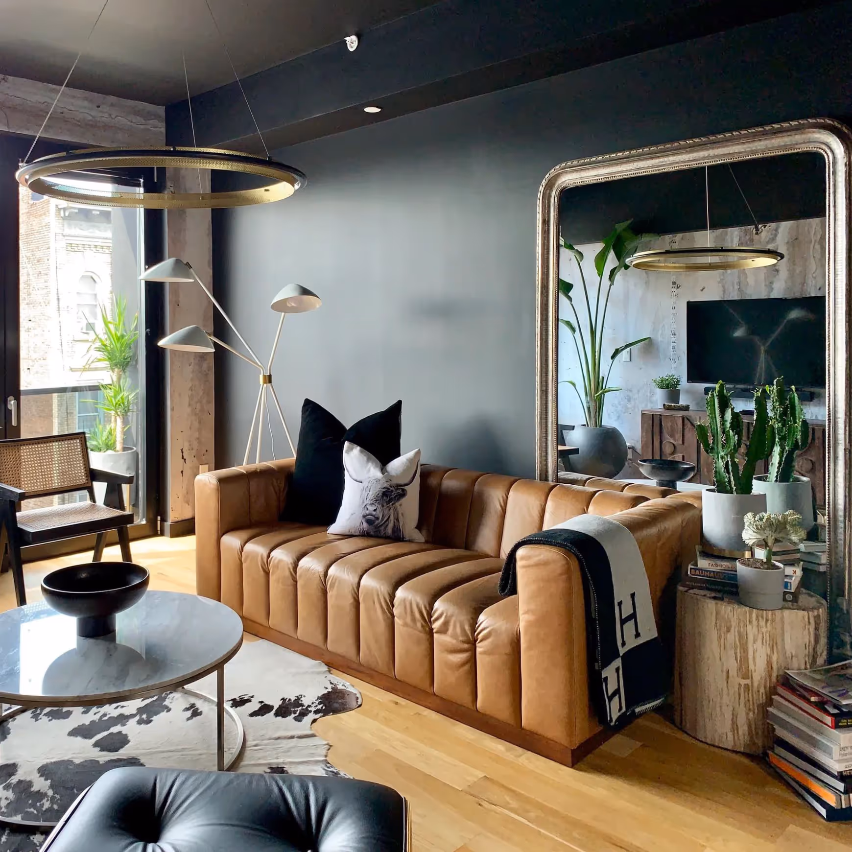 A dark living room with an oversized gold floor standing mirror and a modern tan leather sofa.