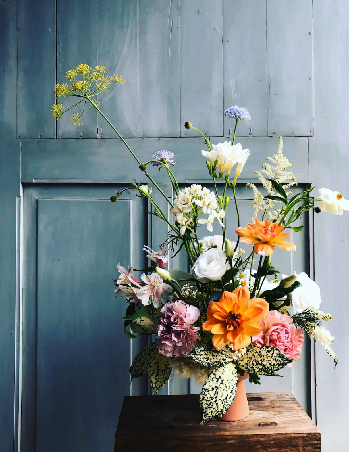 A terracotta vase of colour full summer flowers on a wooden table against an old door painted a distressed ocean blue colour.