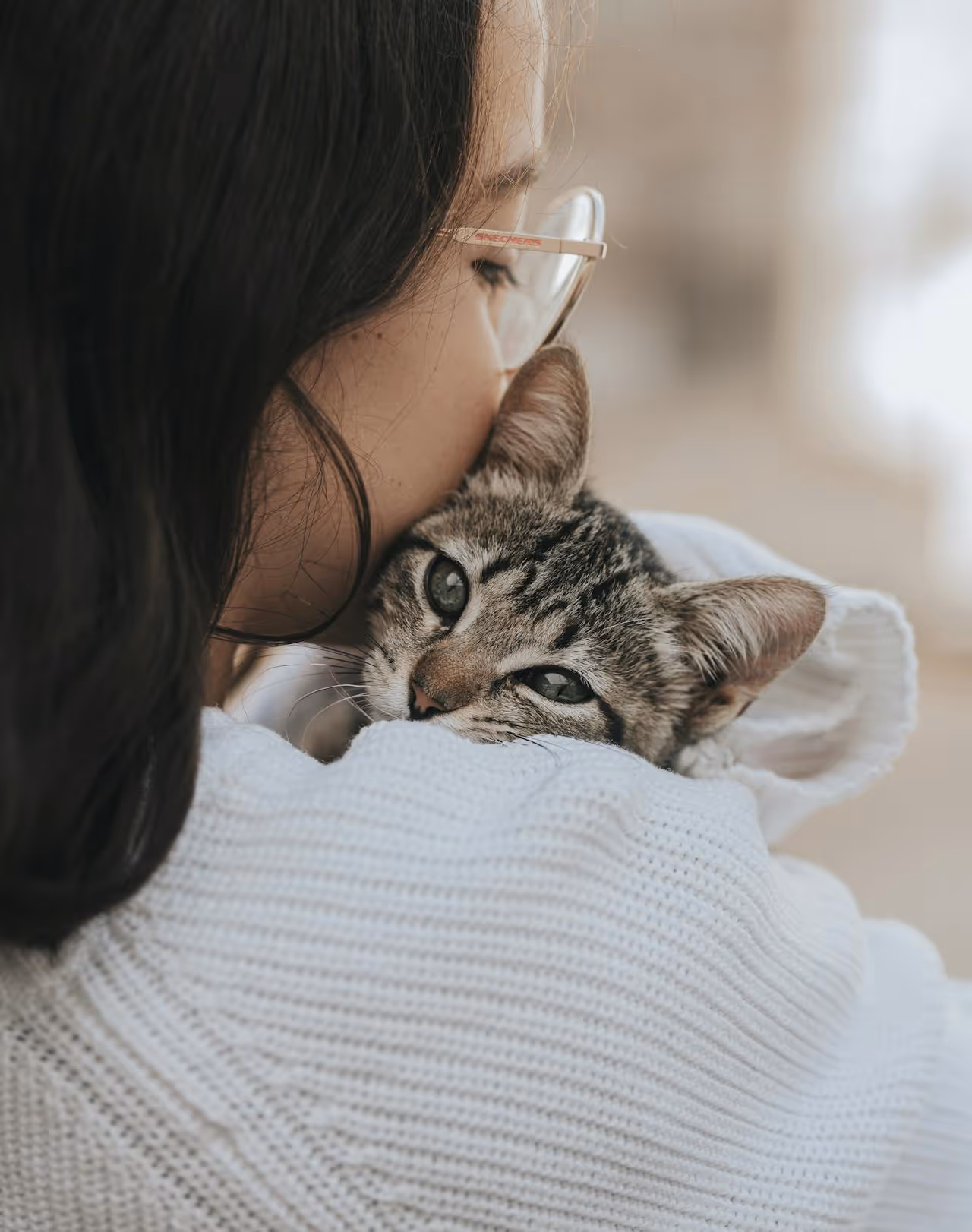 A woman in a white jumper cuddling a tabby kitten.