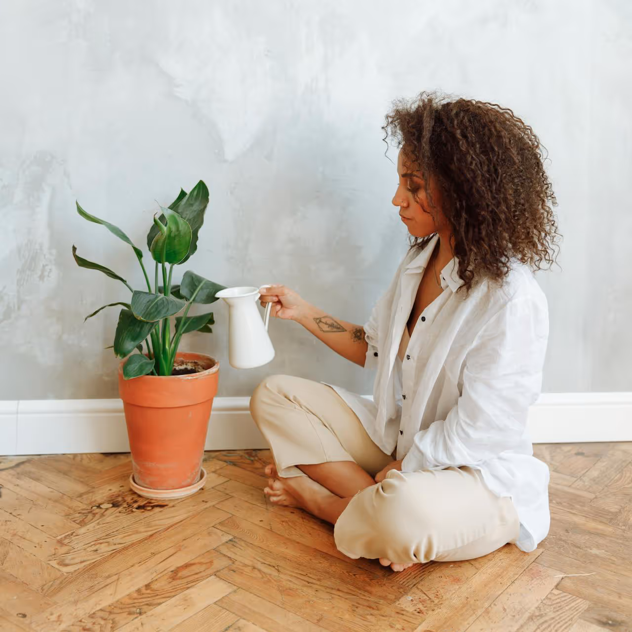 A woman sitting on a wooden floor watering a potted plant.
