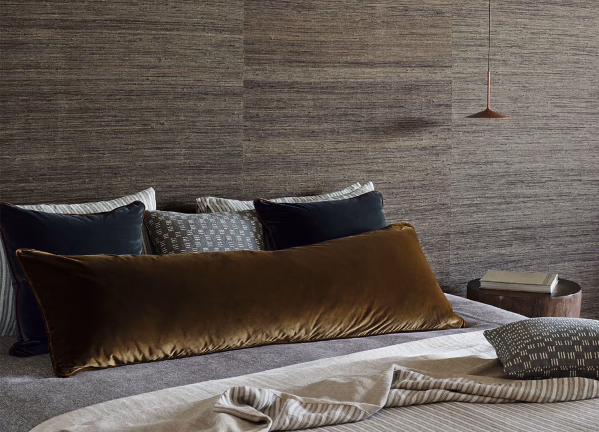 A bed with luxurious mustard and teal velvet cushions and a wall papered with grasscloth.