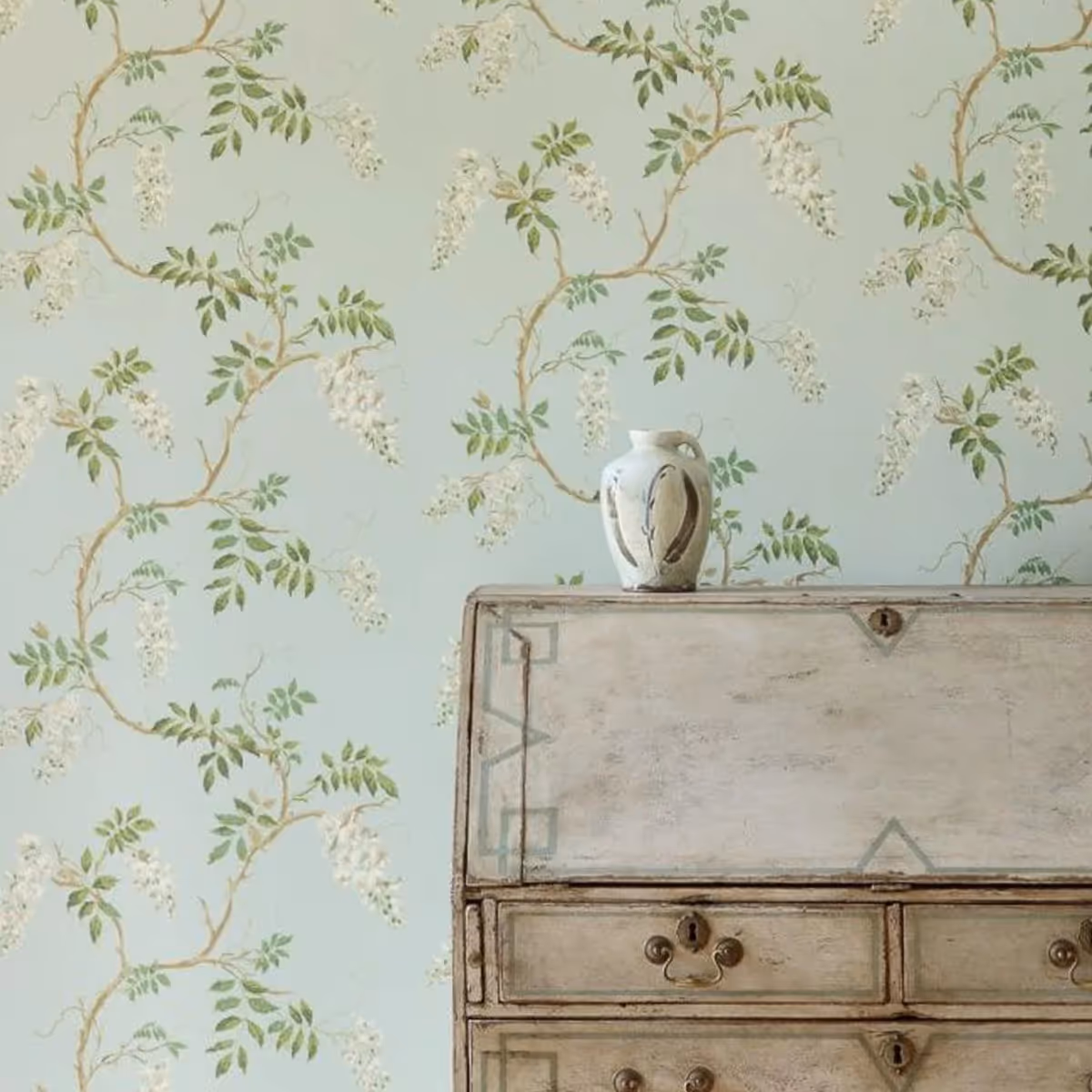 A wooden bureau in front of a wall with a pretty green floral wallpaper