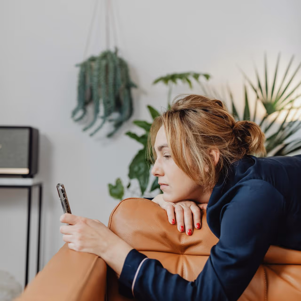 A woman lying on a tan leather sofa staring at her mobile phone.