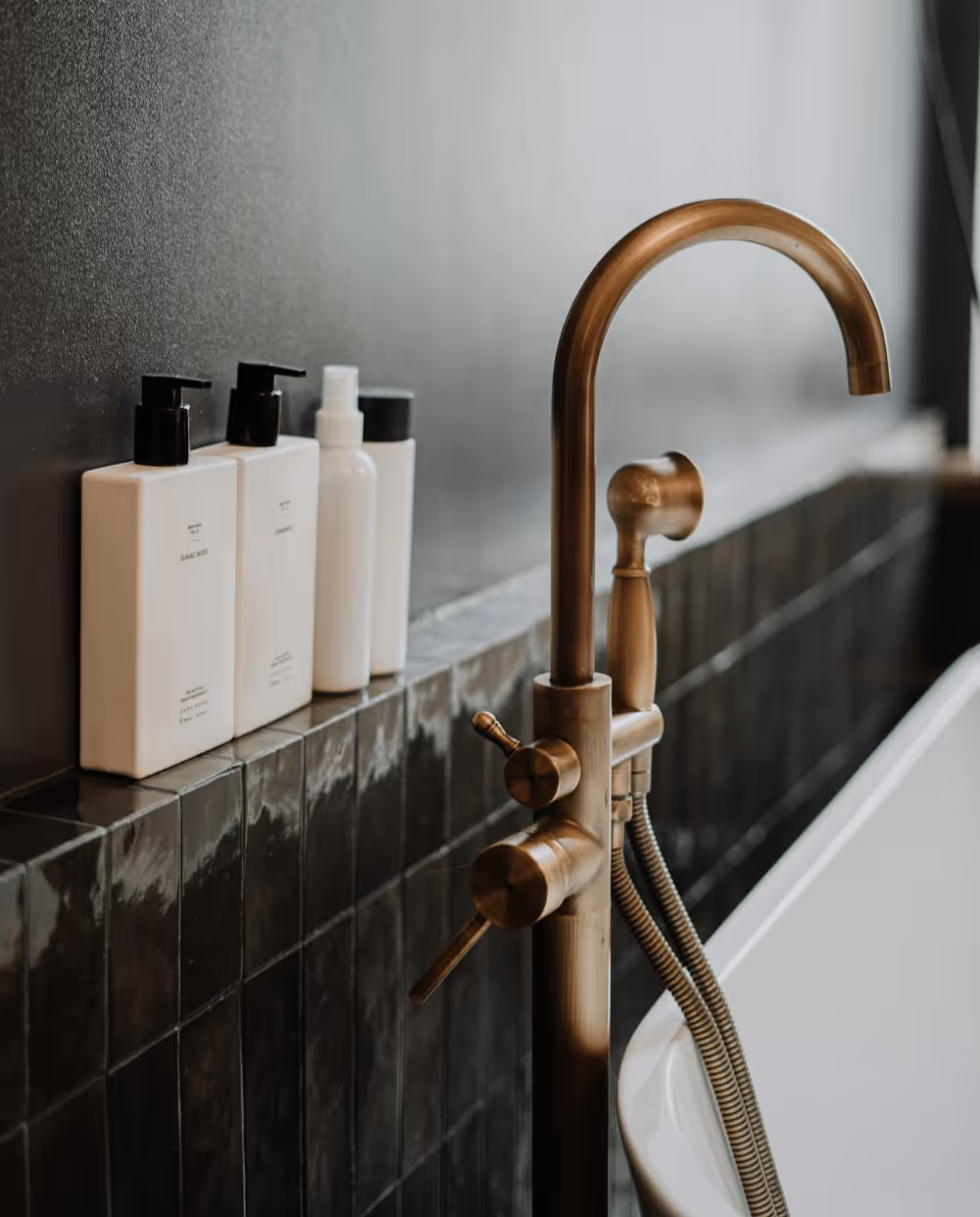 Brass floor mounted bath taps against the backdrop of glossy charcoal tiles