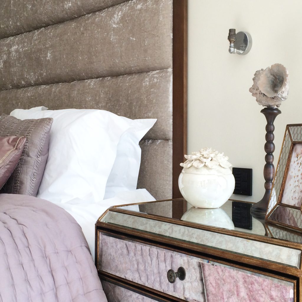 Close up of a bed dressing in white linen and a mauve silk bedspread with a taupe velvet headboard and a walnut and mirrored bedside chest