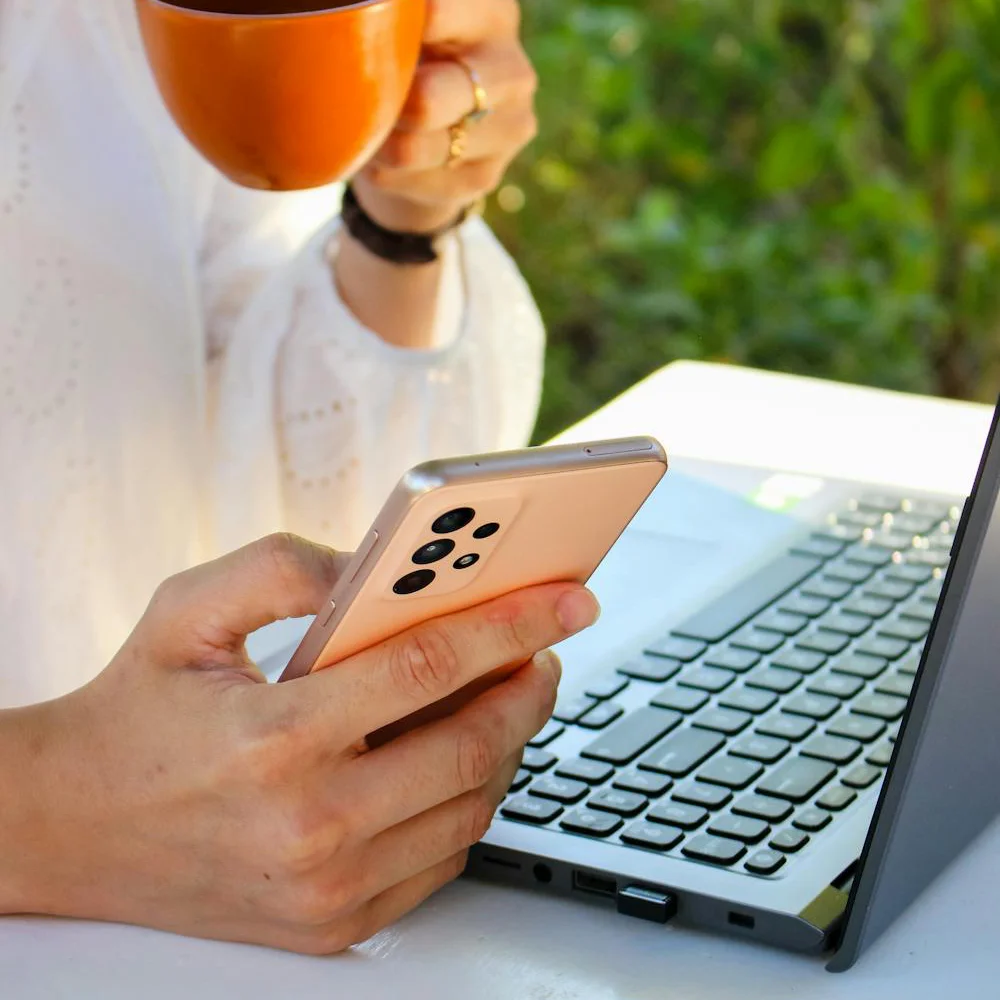 A person drinking a cup of coffee while looking at their phone and laptop